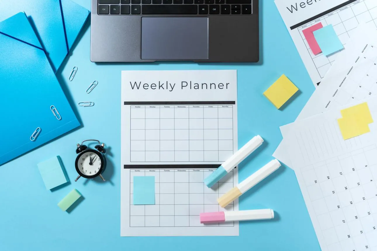 A weekly planner, sticky notes, highlighters and an alarm clock on a blue desk